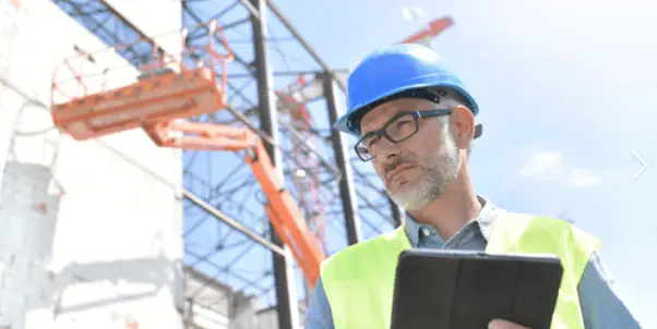 A man wearing a blue hard hat and high-visibility safety vest holds a digital tablet at a construction site.