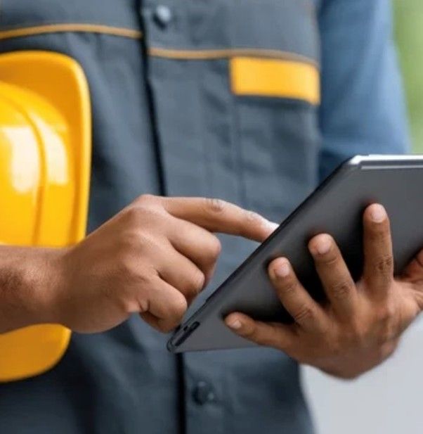 A worker in grey workwear holds a yellow hard hat while using a digital tablet.
