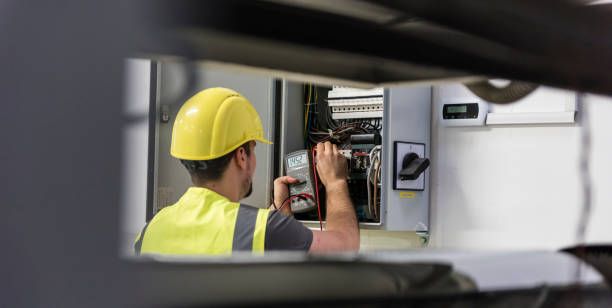 A worker wearing a yellow hard hat and safety vest uses a digital multimeter on an open electrical panel.