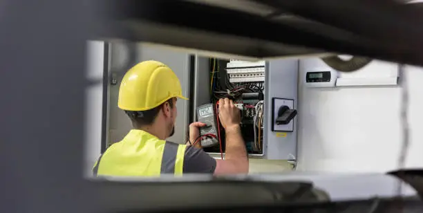 A worker wearing a yellow hard hat and safety vest uses a digital multimeter on an open electrical panel.