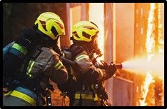 Two firefighters wearing protective gear and bright yellow helmets spray water from a hose at a fire.