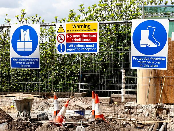 Safety signs on a fence mandate high-visibility clothing, protective footwear, and warn of a multi-hazard area.
