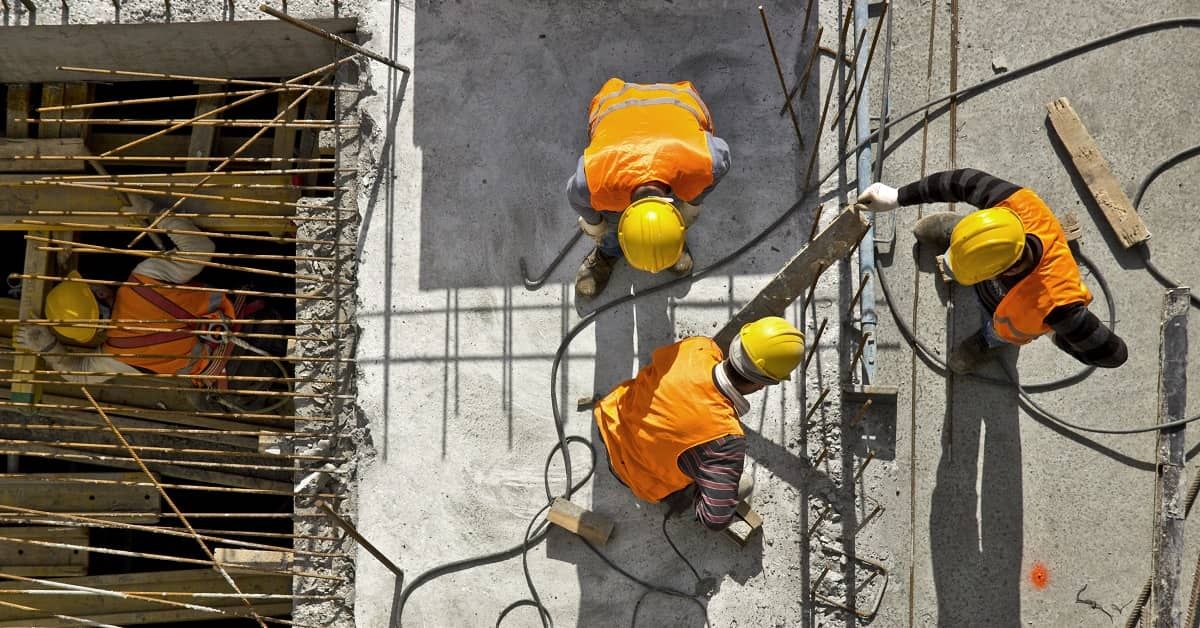 Four construction workers wearing yellow hard hats and orange safety vests work on a concrete structure with exposed rebar.