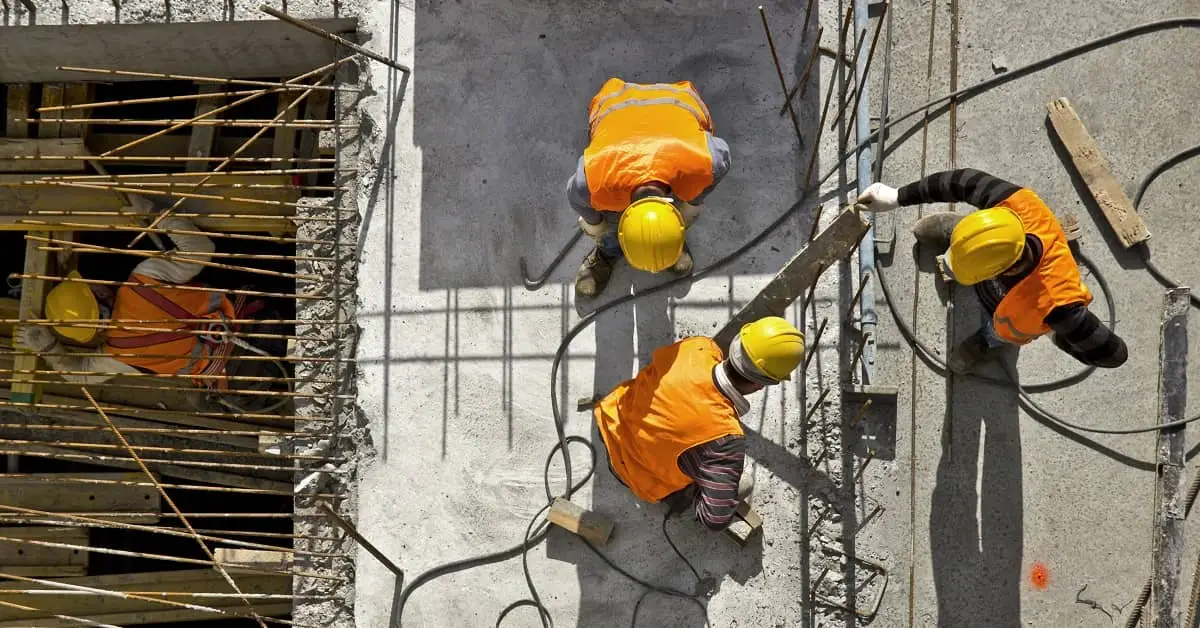 Four construction workers wearing yellow hard hats and orange safety vests work on a concrete structure with exposed rebar.