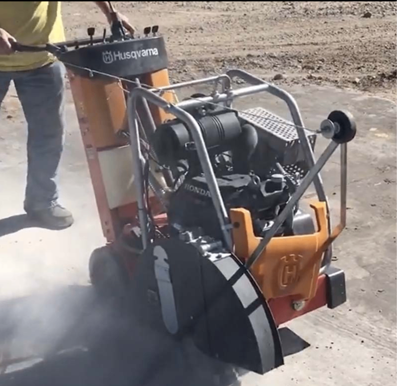 A worker operates a walk-behind Husqvarna concrete saw, creating a thick cloud of dust while cutting concrete.
